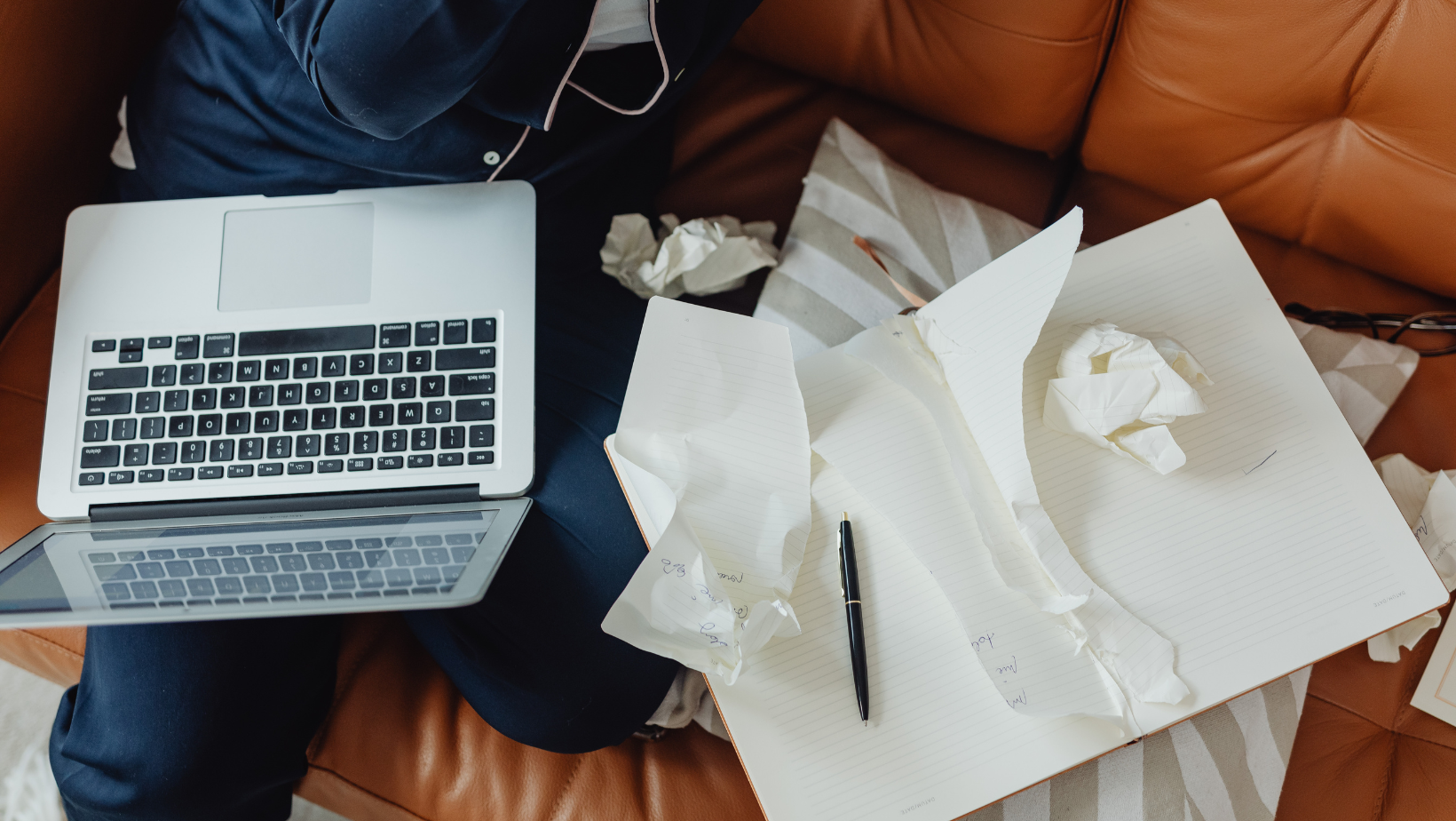 Writer sitting on a couch with a laptop and notebook surrounded by crumpled paper, symbolizing writer’s block and the struggle to start writing.