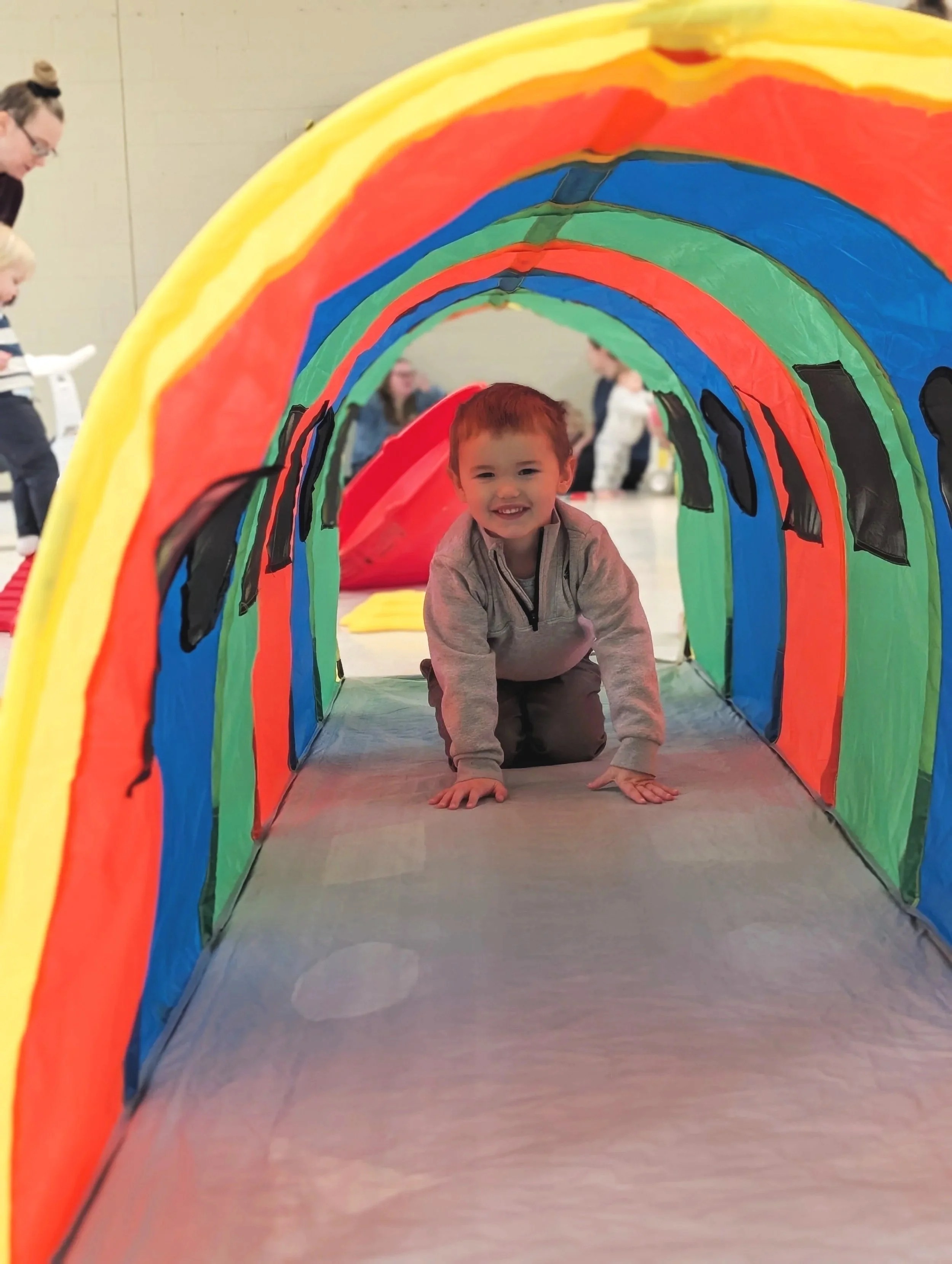 A young boy crawling through a colorful fabric tunnel with rainbow stripes at an indoor play area. He is smiling and wearing a gray jacket.