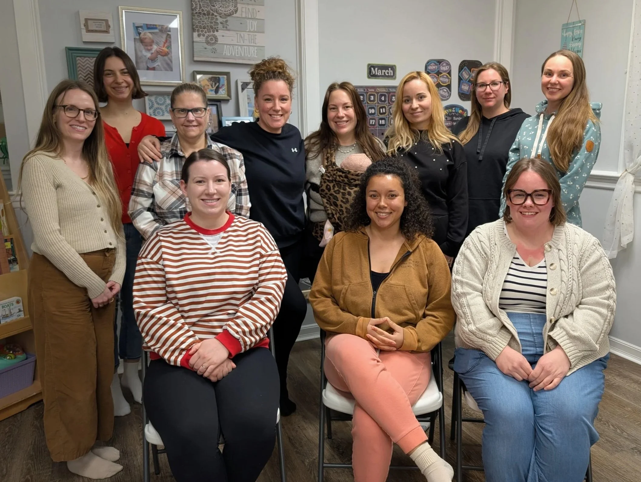 Group of twelve women posing indoors in a casual setting, some sitting and some standing, smiling at the camera.