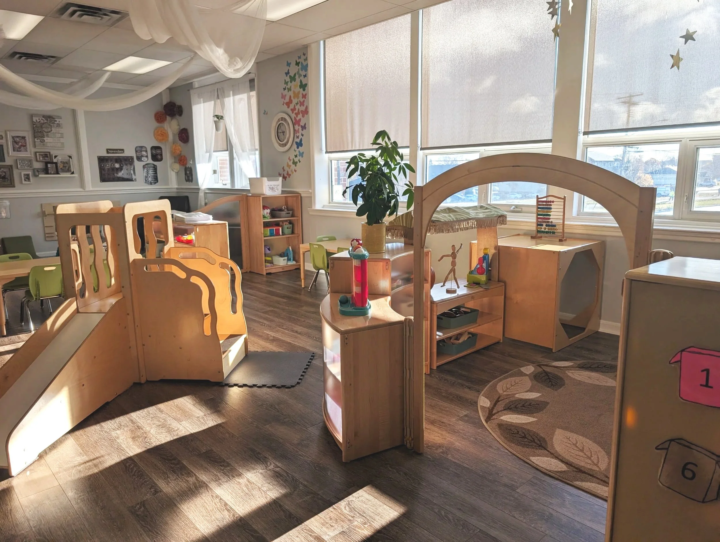 Brightly lit preschool classroom with wooden play structures, toys, small tables and chairs, wall decorations, and large windows with sunlight streaming in.