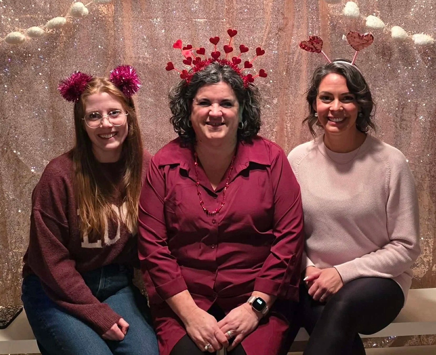 Three women sitting together against a glittery backdrop, wearing festive headbands with hearts and pom-poms, smiling for a photo.