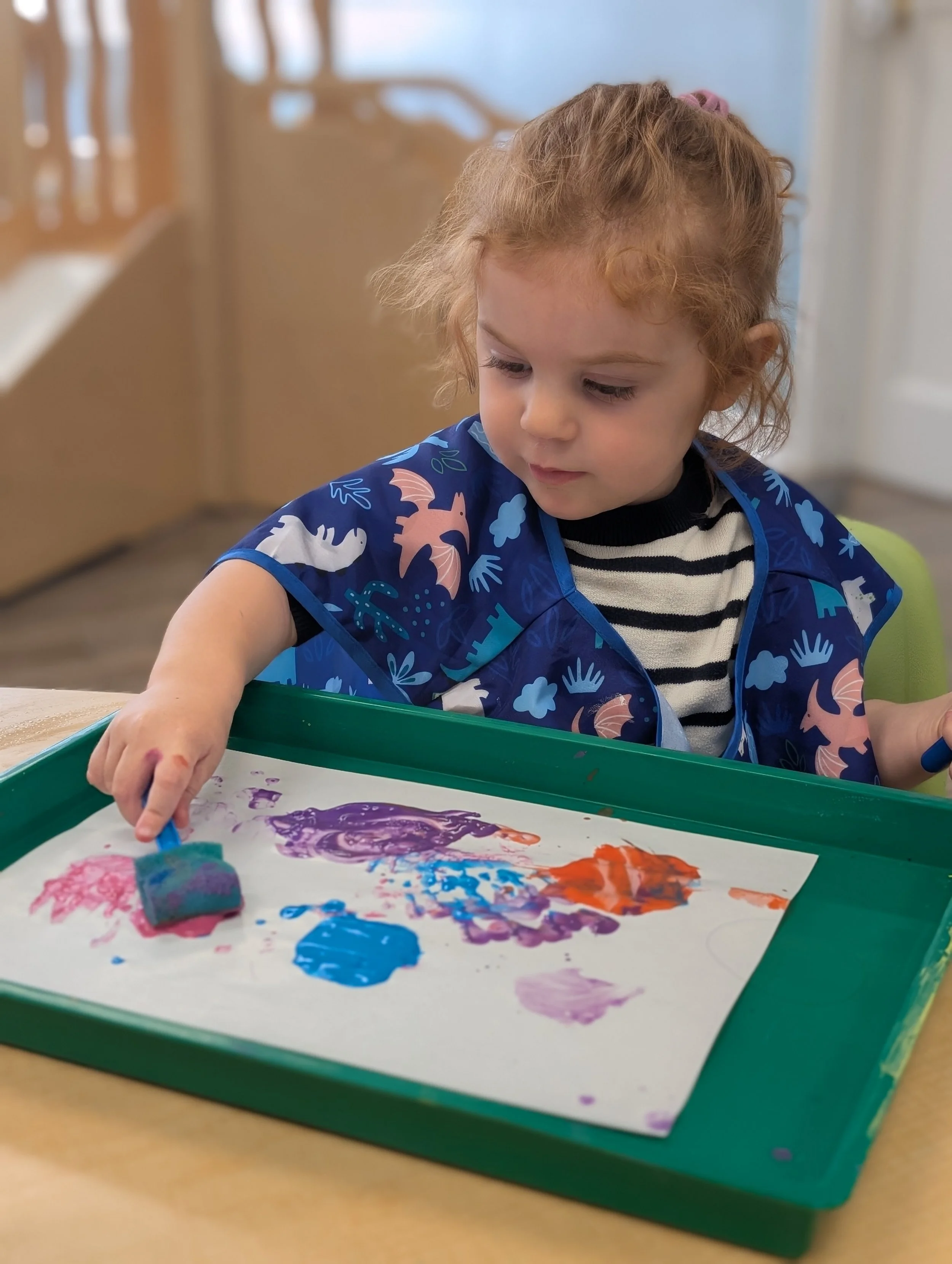 A young girl with curly red hair sitting at a table, using a foam brush to paint on a piece of paper with colorful paint, wearing a blue apron with dinosaur and cloud patterns.