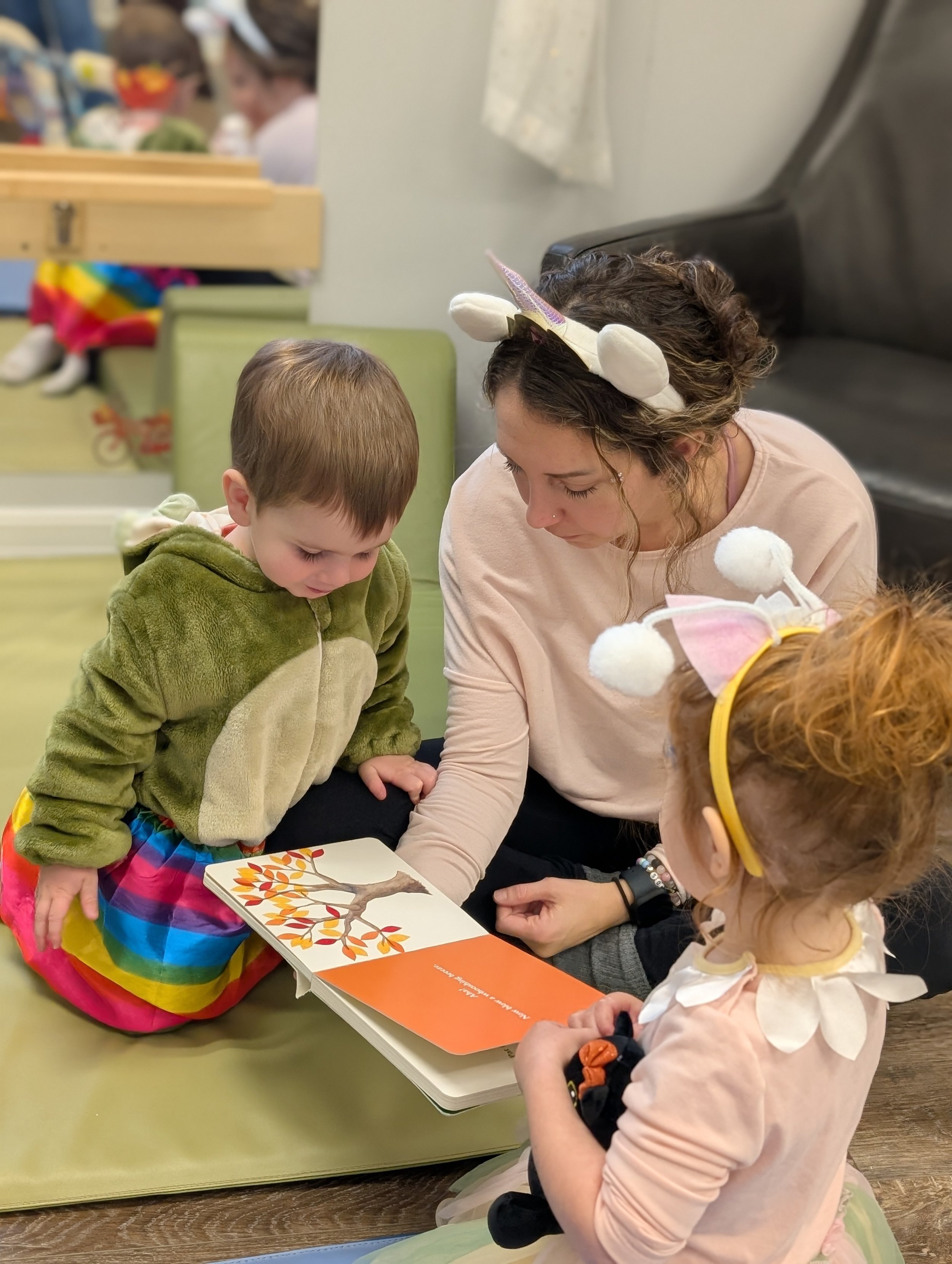 A woman and two children wearing unicorn headbands are reading a picture book together on the floor in a cozy indoor setting.