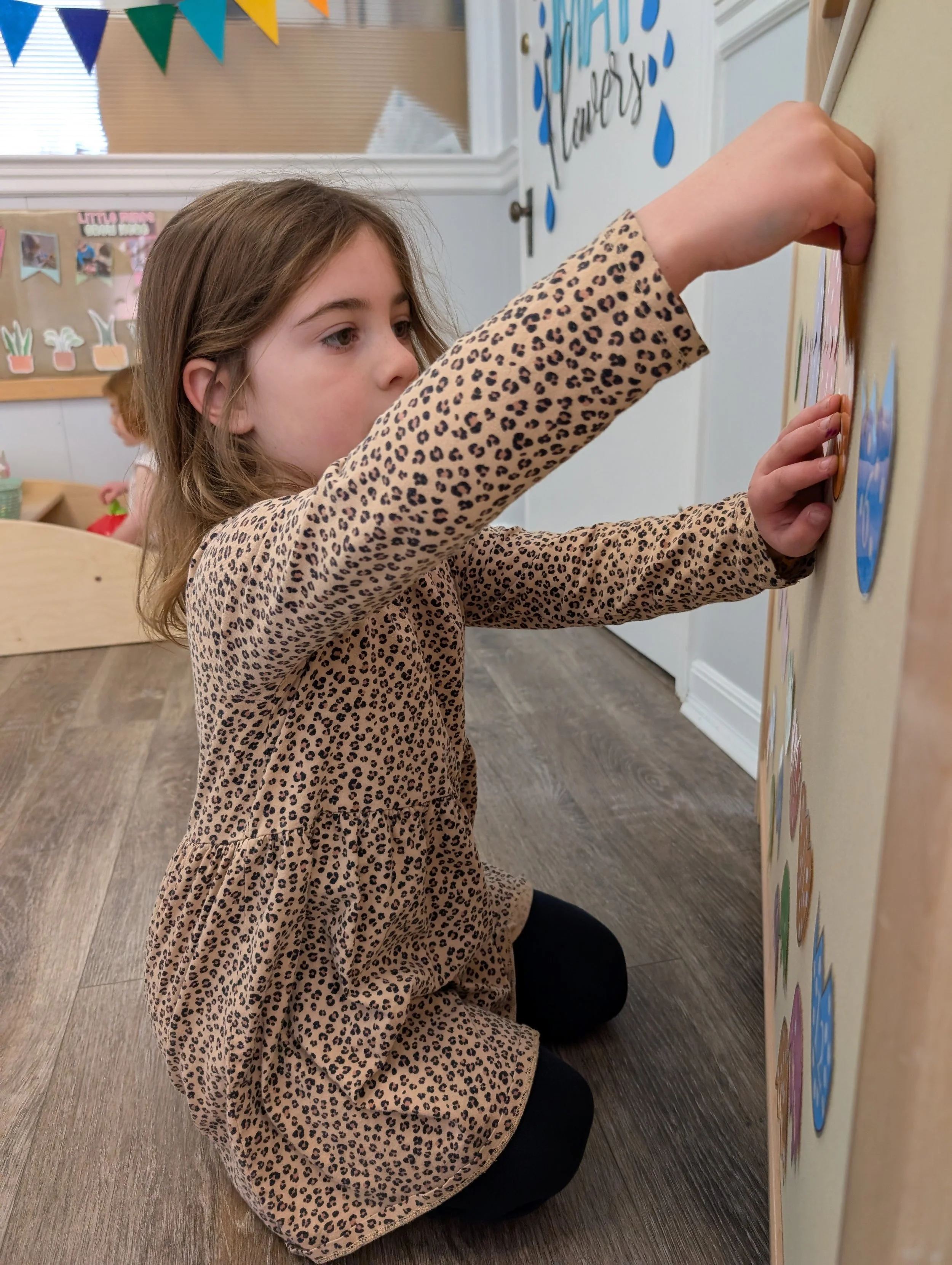 A young girl with long hair, dressed in a leopard-print dress, kneeling on the floor and placing stickers on a display board in a classroom.