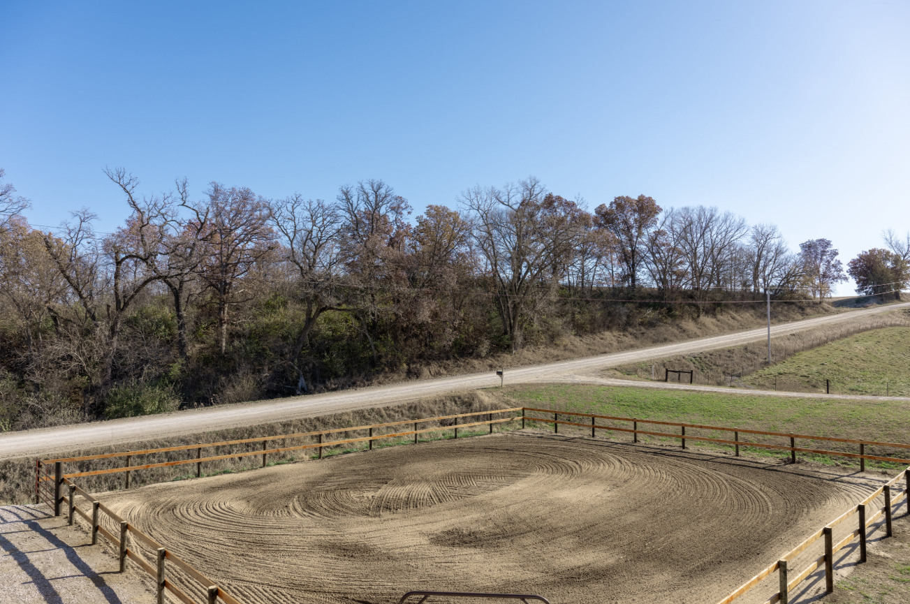 An outdoor riding arena with a dirt surface, surrounded by a wooden fence, and a rural landscape with leafless trees, a dirt road, and hills under a clear blue sky.