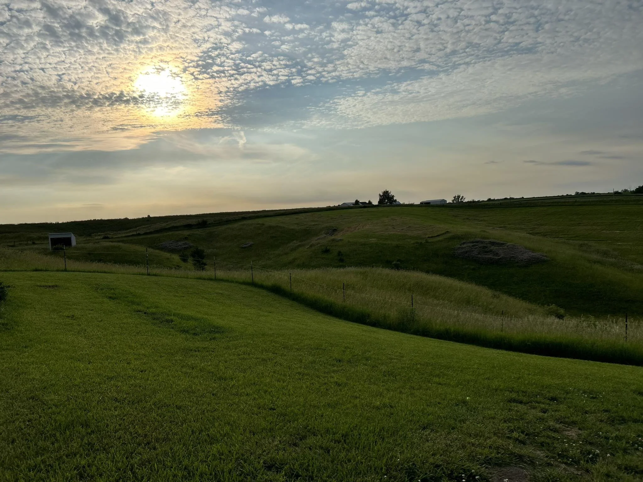 A scenic view of green rolling hills with a partly cloudy sky and the sun setting or rising in the background.