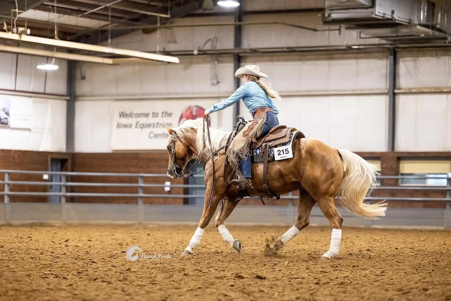A young woman riding a palomino horse in an indoor arena, wearing a cowboy hat, a denim shirt, and western attire, with the horse practicing riding skills.