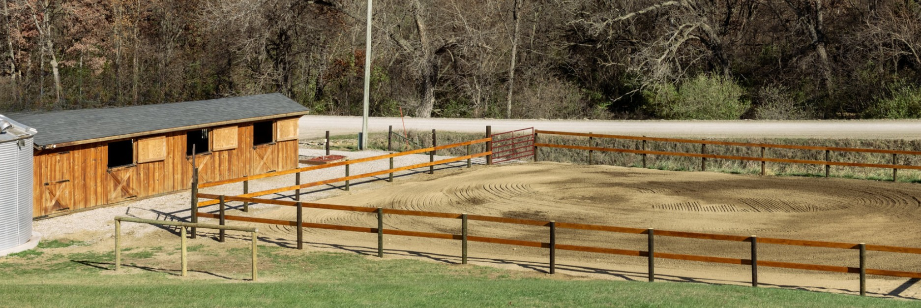 Empty outdoor horse riding arena with wooden fence, a wooden shed, and a gravel road with trees in the background.
