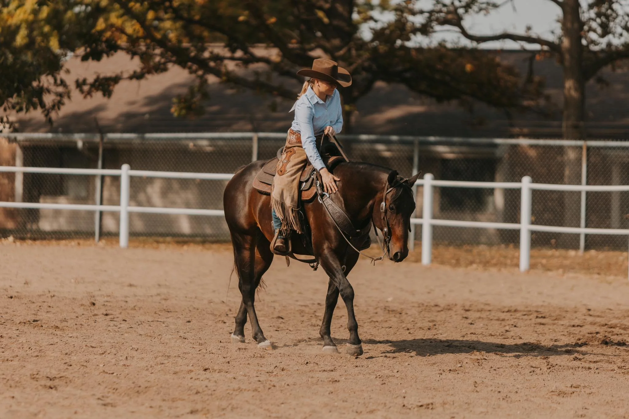 A woman wearing a cowboy hat, blue shirt, and jeans riding a brown horse on an outdoor dirt arena, with trees and a fence in the background.