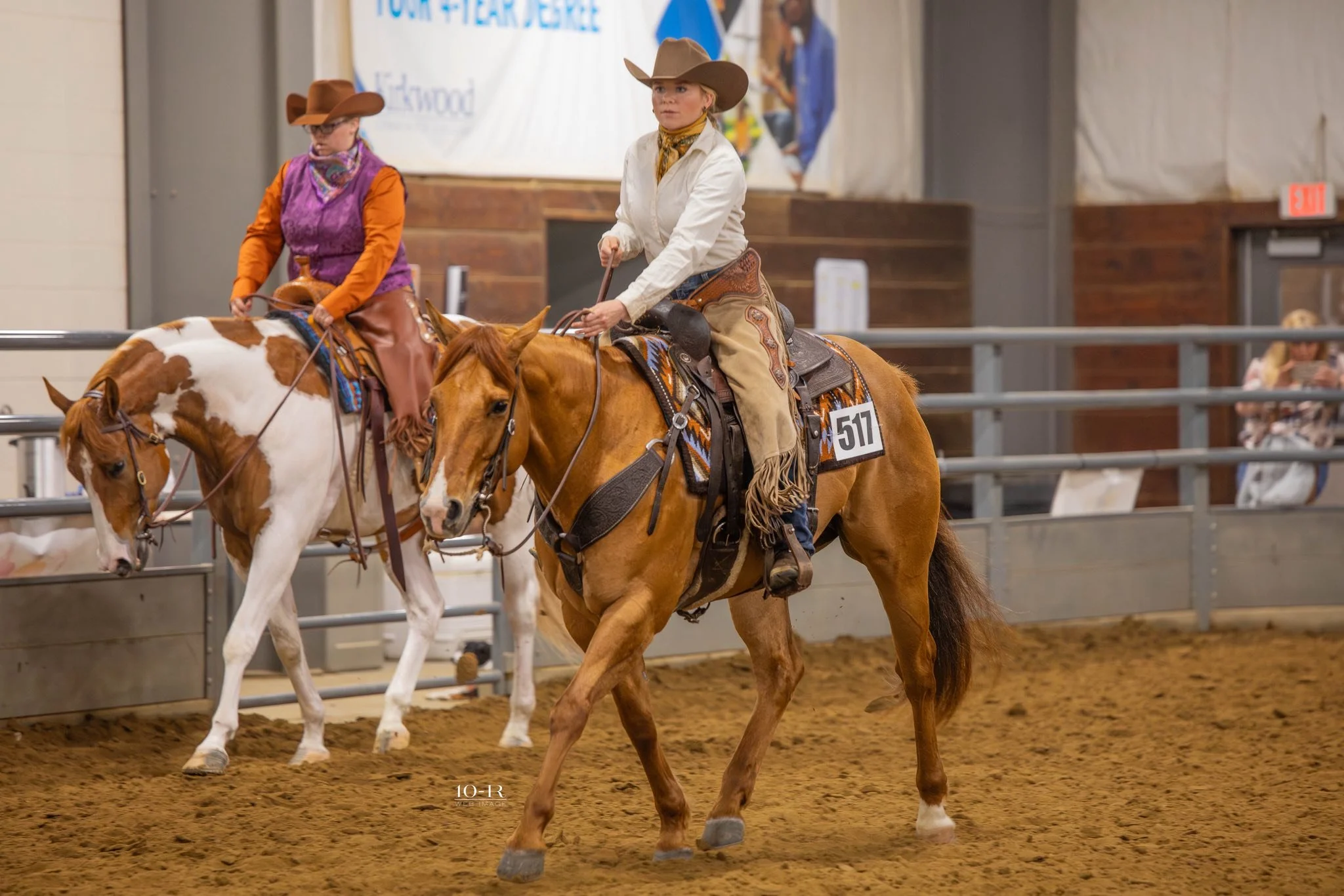 Two women wearing cowboy hats and western attire riding horses in an indoor arena at a rodeo or horse show event.