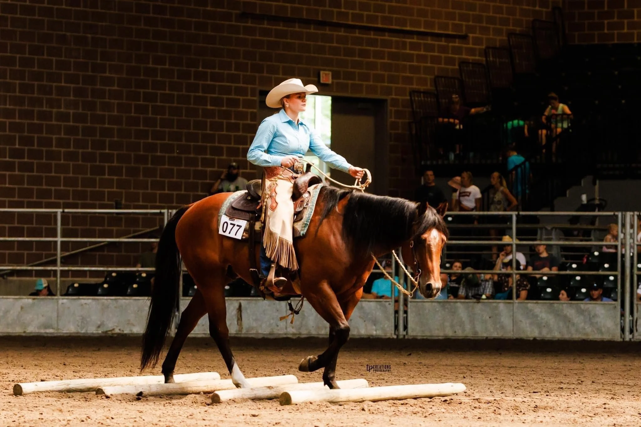 A woman riding a horse during a Western riding event in an indoor arena, with spectators watching from the stands.