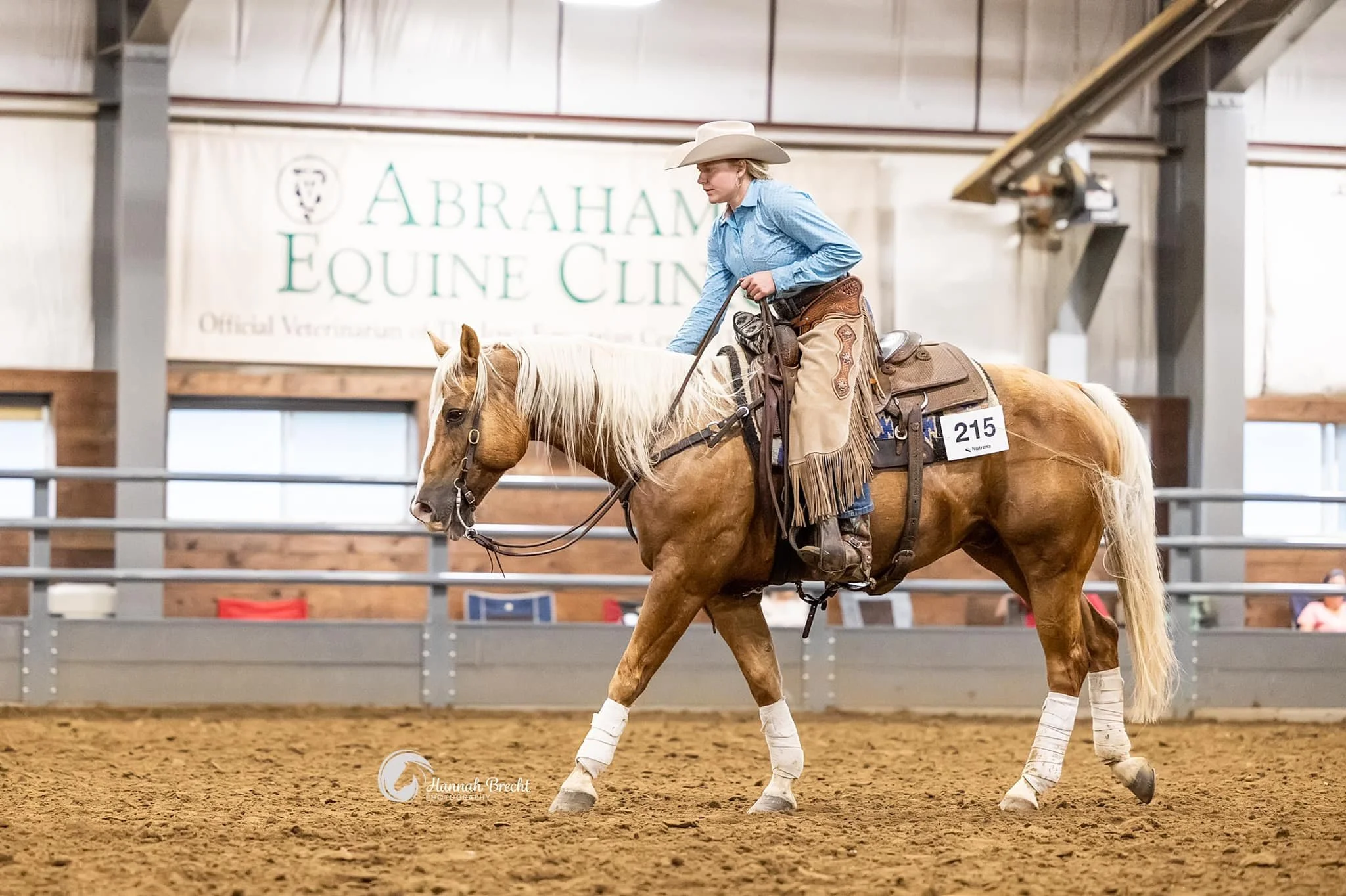 A woman in cowboy attire riding a palomino horse inside an indoor arena. The woman is wearing a cowboy hat, blue shirt, chaps, and riding boots. The horse has white leg wraps and a saddle with a number 215 attached. There is a sign in the background that reads 'Abraham Equine Clinic'.