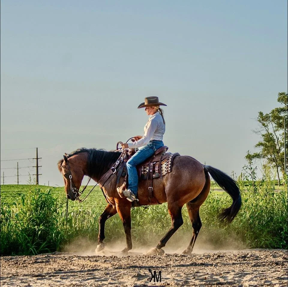 A woman wearing a cowboy hat, white long-sleeve shirt, and blue jeans riding a brown horse on a dirt path with green grass and trees in the background under a clear blue sky.