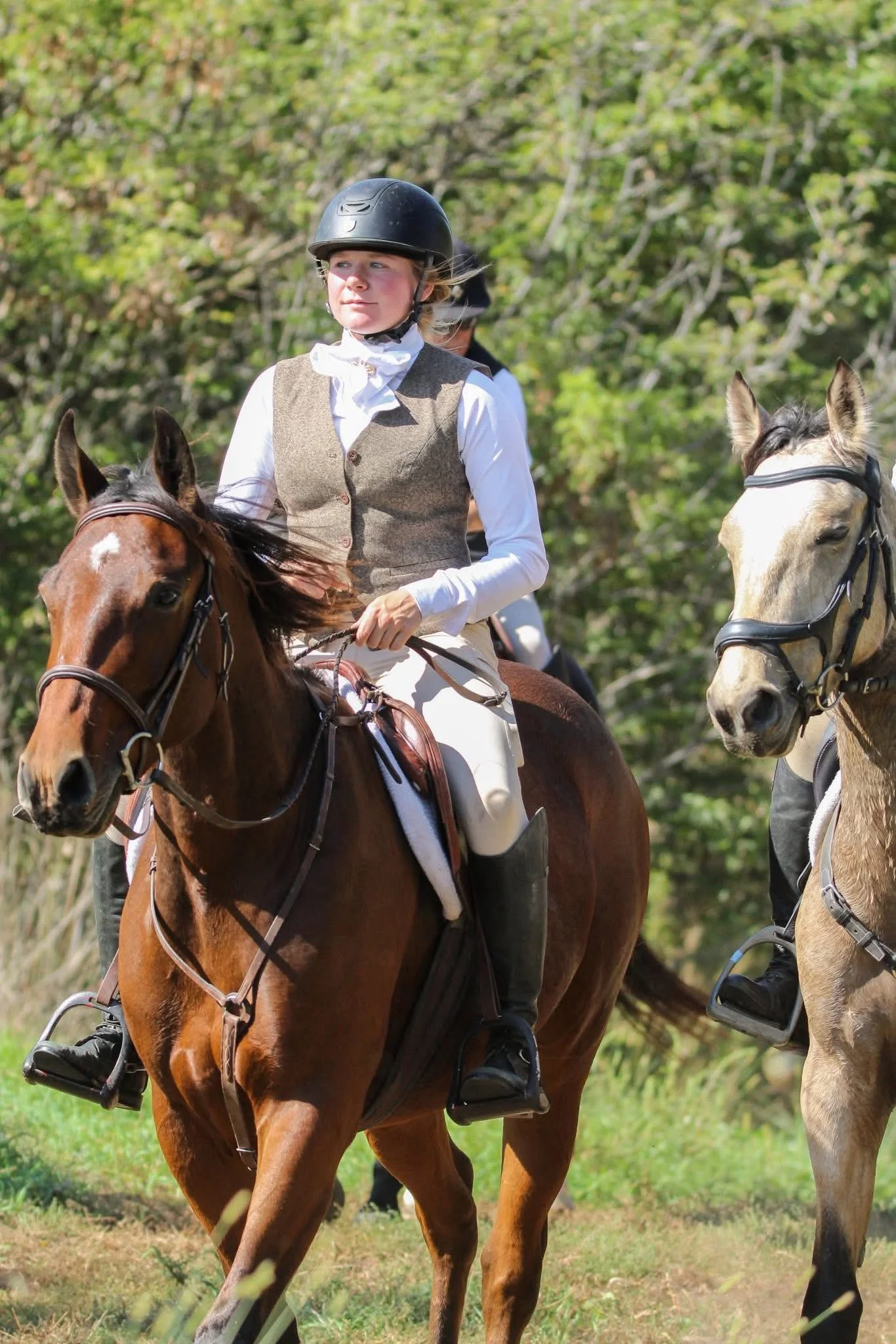 A woman wearing a helmet and vintage riding attire riding a brown horse outdoors, with another rider and horse visible in the background, surrounded by green trees.