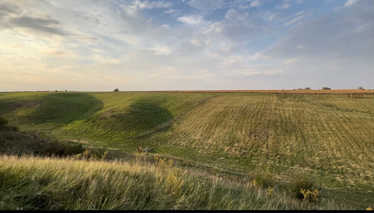 A vast open landscape with rolling grassy hills and cultivated fields under a partly cloudy sky.