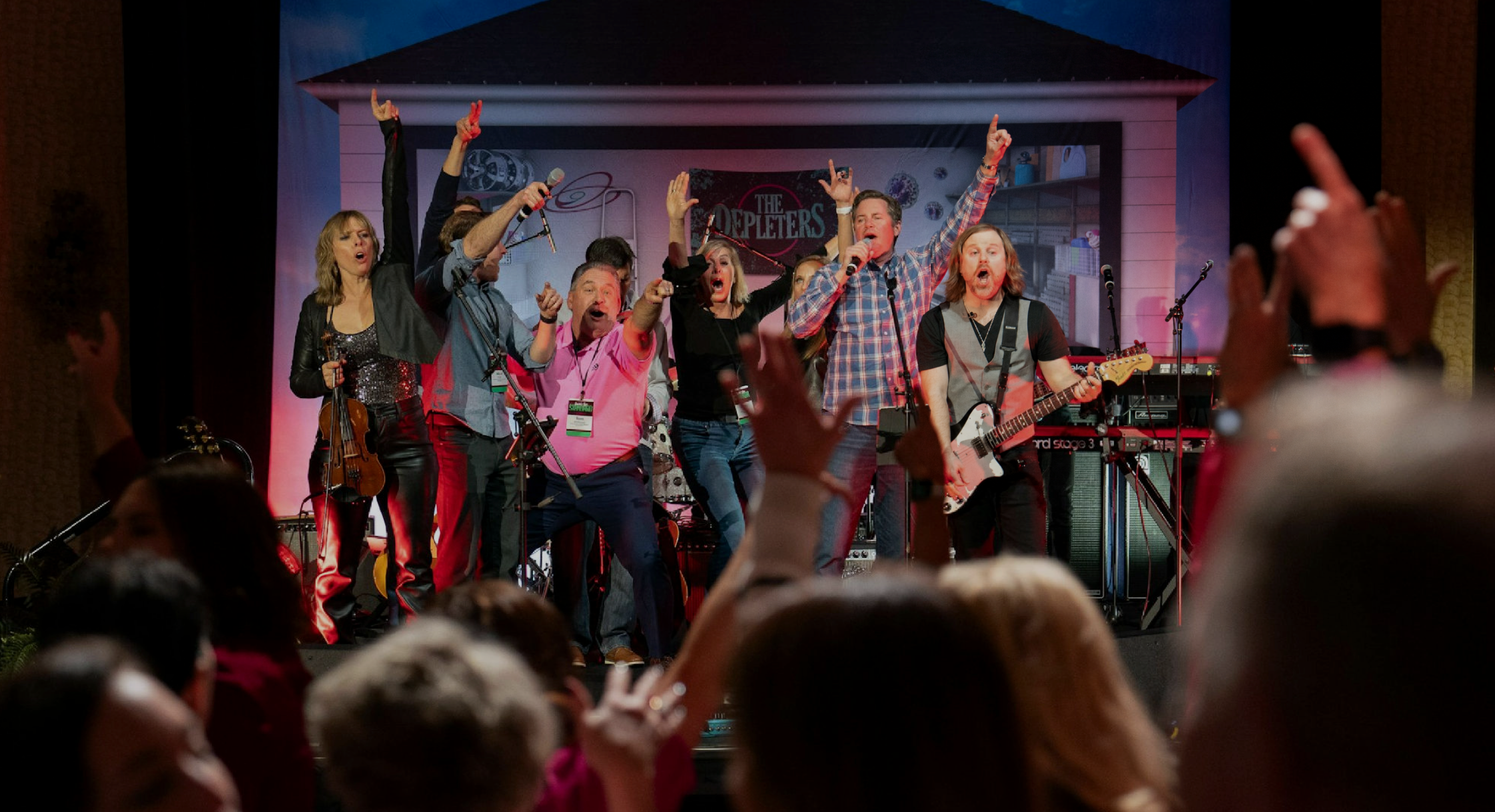 Group of performers singing and playing musical instruments on a stage during a concert, with a sign reading 'The Detectives' in the background.
