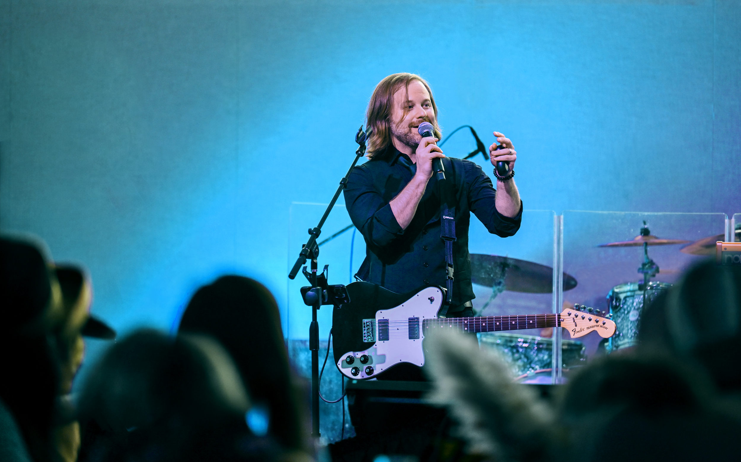 A man with shoulder-length hair and a beard singing into a microphone on stage while holding an electric guitar. The background is illuminated with blue lighting and a drum set is partially visible behind him.
