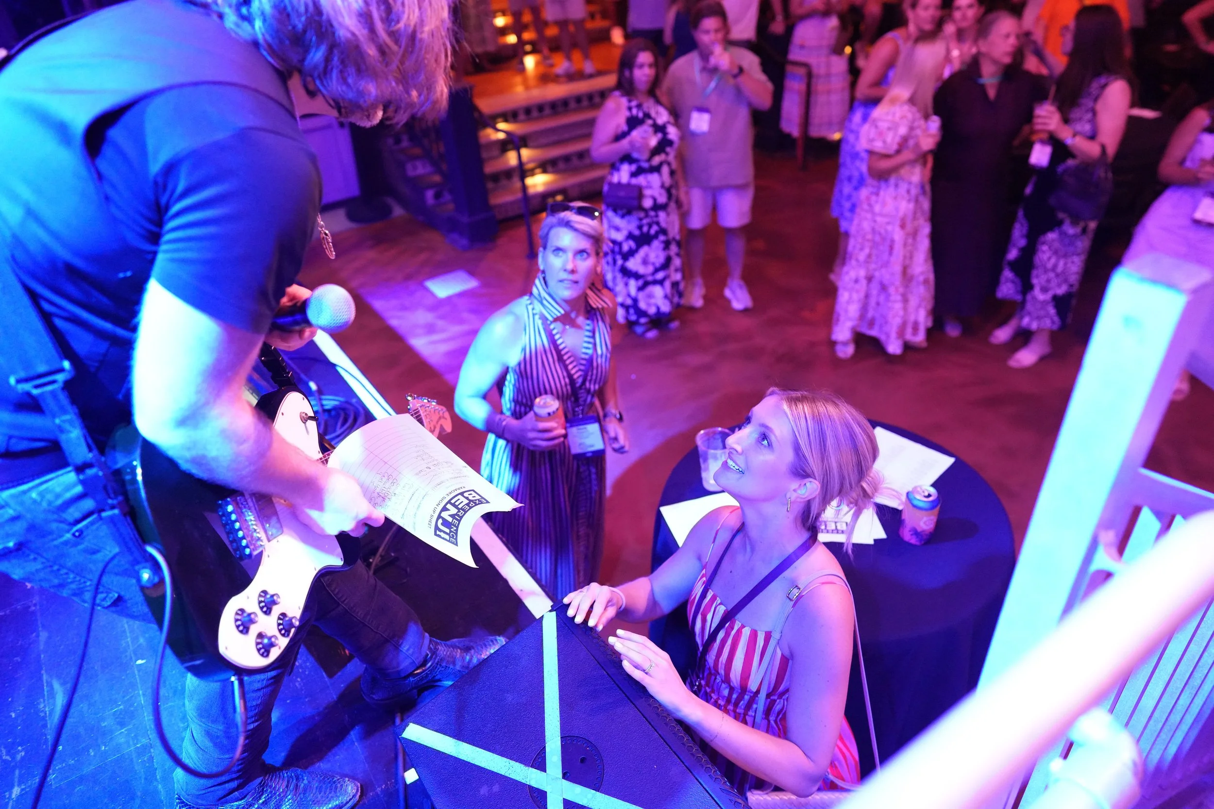 A woman in a colorful dress signs a book at a social gathering. She is seated at a table with papers, and several people stand around her, some holding drinks. The setting has dim colorful lighting and a lively atmosphere.