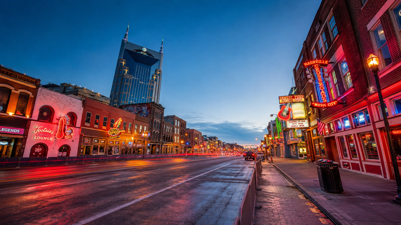 Downtown Nashville street scene at dusk with neon signs, historic buildings, and the AT&T building in the background.