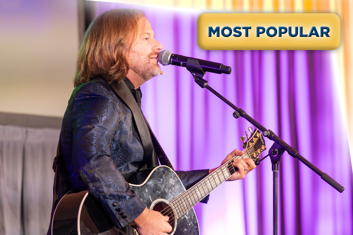 A man with shoulder-length hair and a beard, wearing a dark patterned jacket, is singing into a microphone while playing an acoustic guitar on stage with purple curtains in the background. A yellow label with blue text says "MOST POPULAR" in the top right corner.