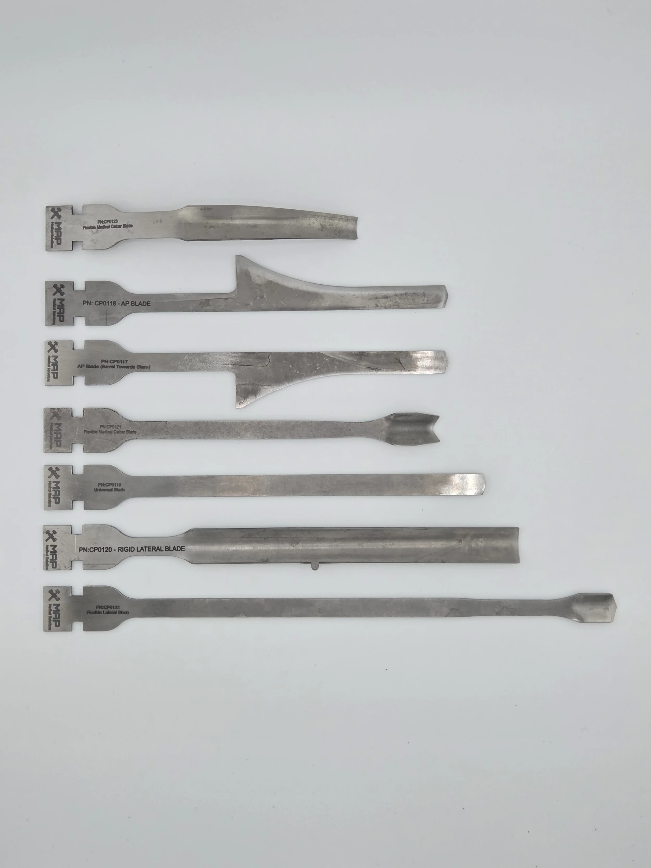 A set of six metal dental or craft blades of varying shapes, sizes, and types, arranged in a vertical line on a white background.