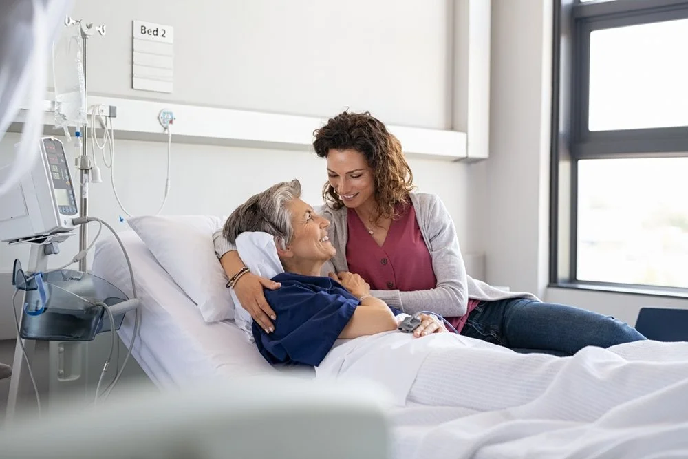 A woman sitting beside a hospital bed, smiling and holding an elderly woman who is lying in the bed. The elderly woman appears to be a patient, wearing hospital attire, with medical equipment nearby. The room is bright with a large window.