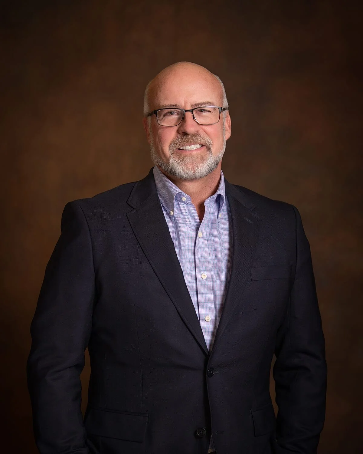 Portrait of a middle-aged man with glasses, a beard, and bald head, wearing a dark blazer and a light-colored button-up shirt, standing against a brown gradient background.