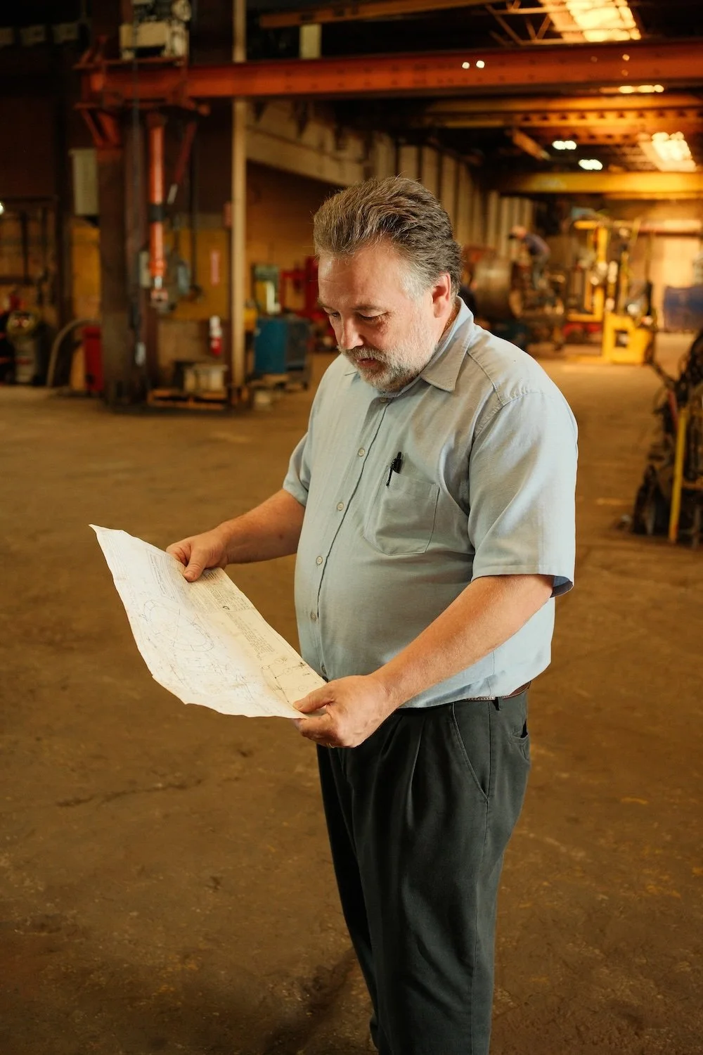 A middle-aged man with gray hair and beard wearing a light gray shirt and black pants, standing in a large industrial warehouse, holding and reading a large technical blueprint or schematic.