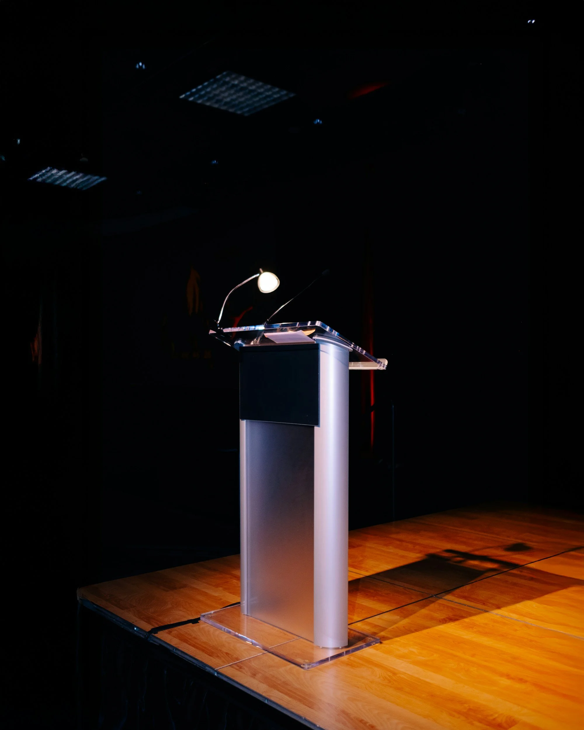 Empty podium with a microphone and a light on it, set on a stage with a wooden floor, in a dark room.