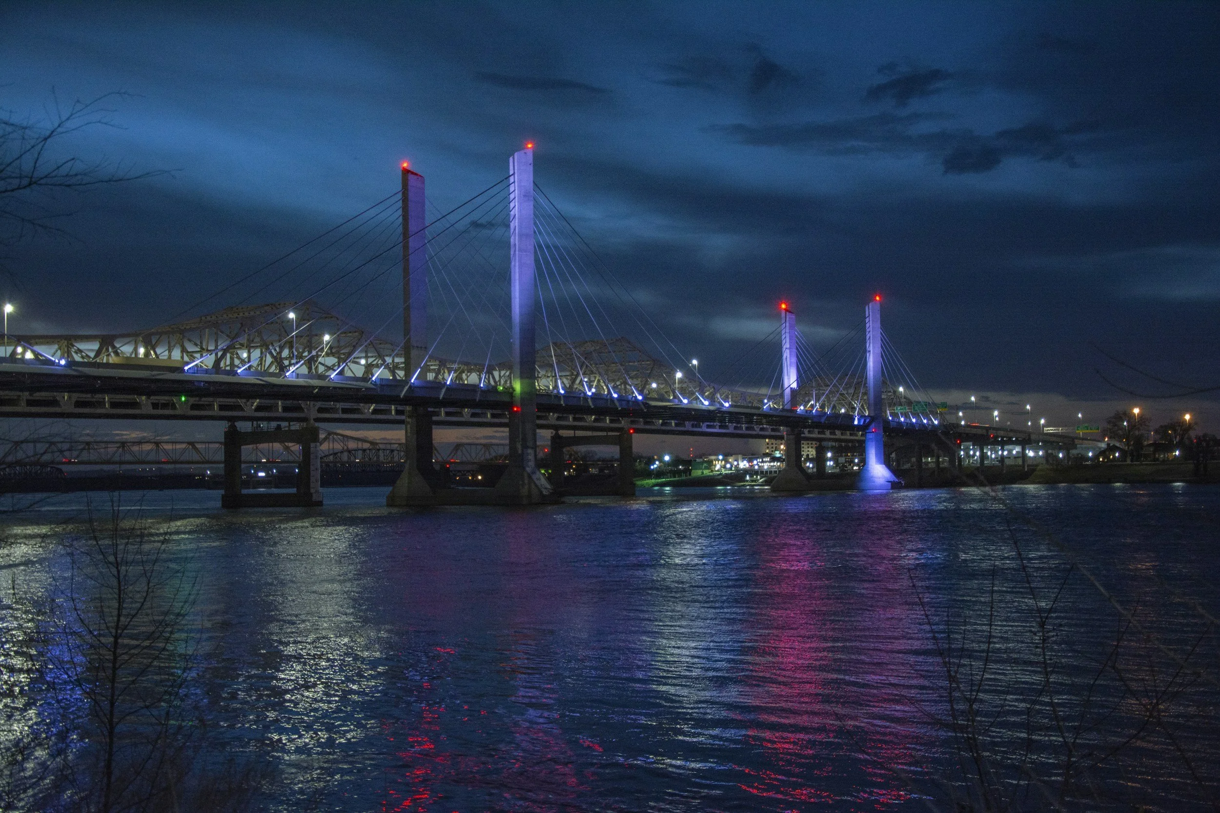 Nighttime view of a bridge illuminated with colorful lights over a river, with reflections on the water and a dark, cloudy sky.
