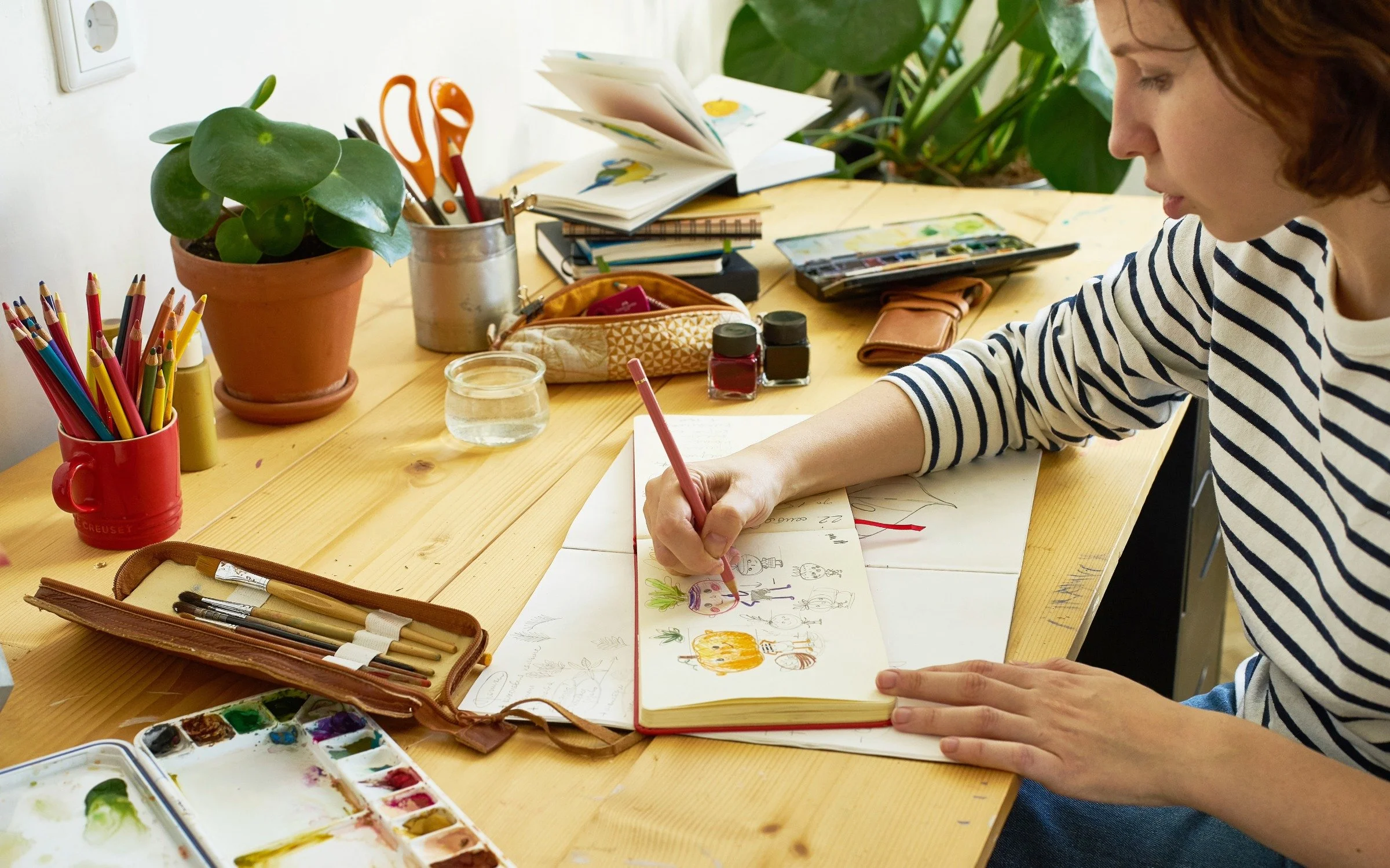 A woman sitting at a wooden desk drawing in a sketchbook with colored pencils. The desk has art supplies, plants, water, and notebooks.