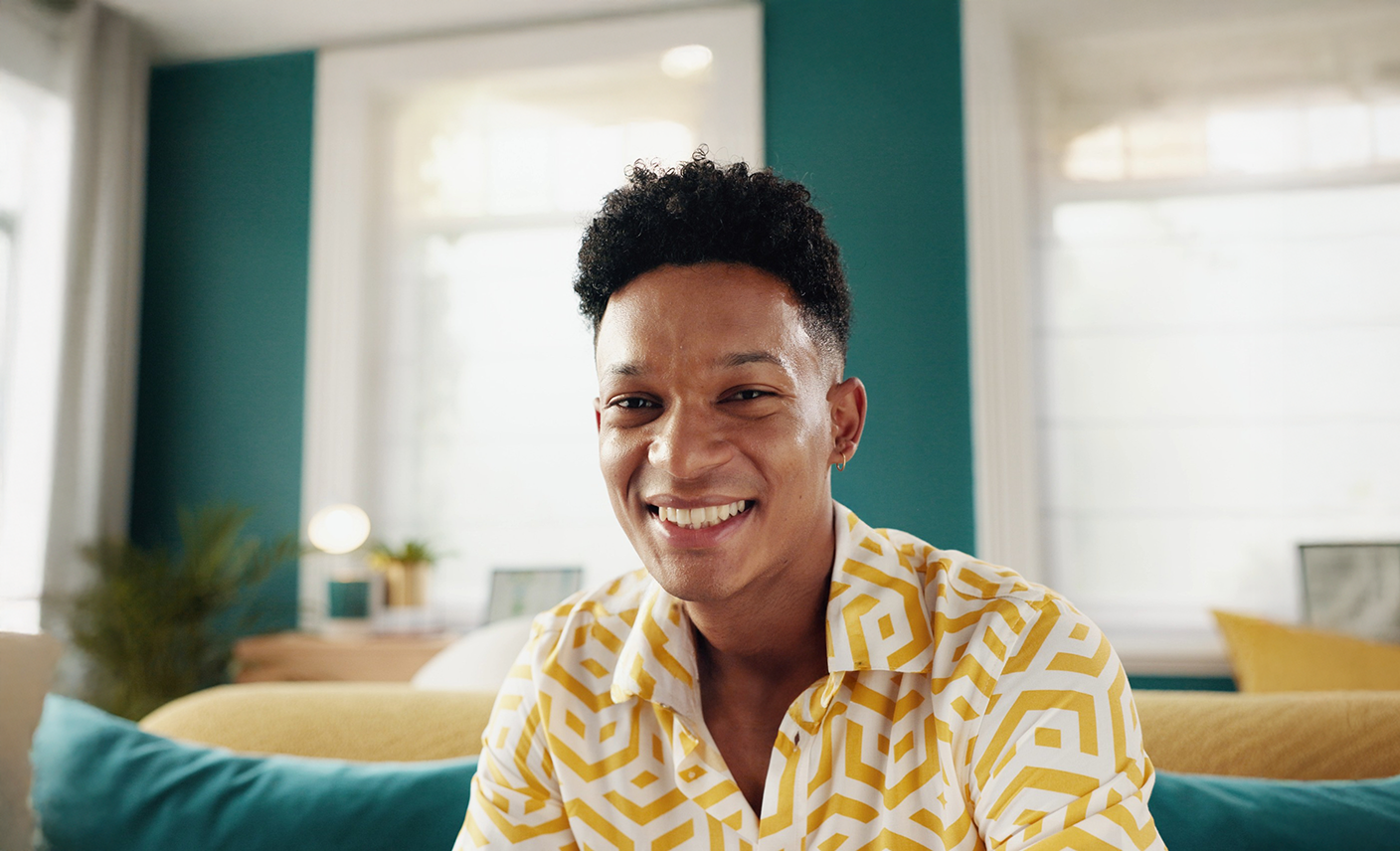 A young man with short curly black hair, smiling, wearing a yellow and white patterned shirt, sitting in a brightly lit living room with teal walls and white window blinds.
