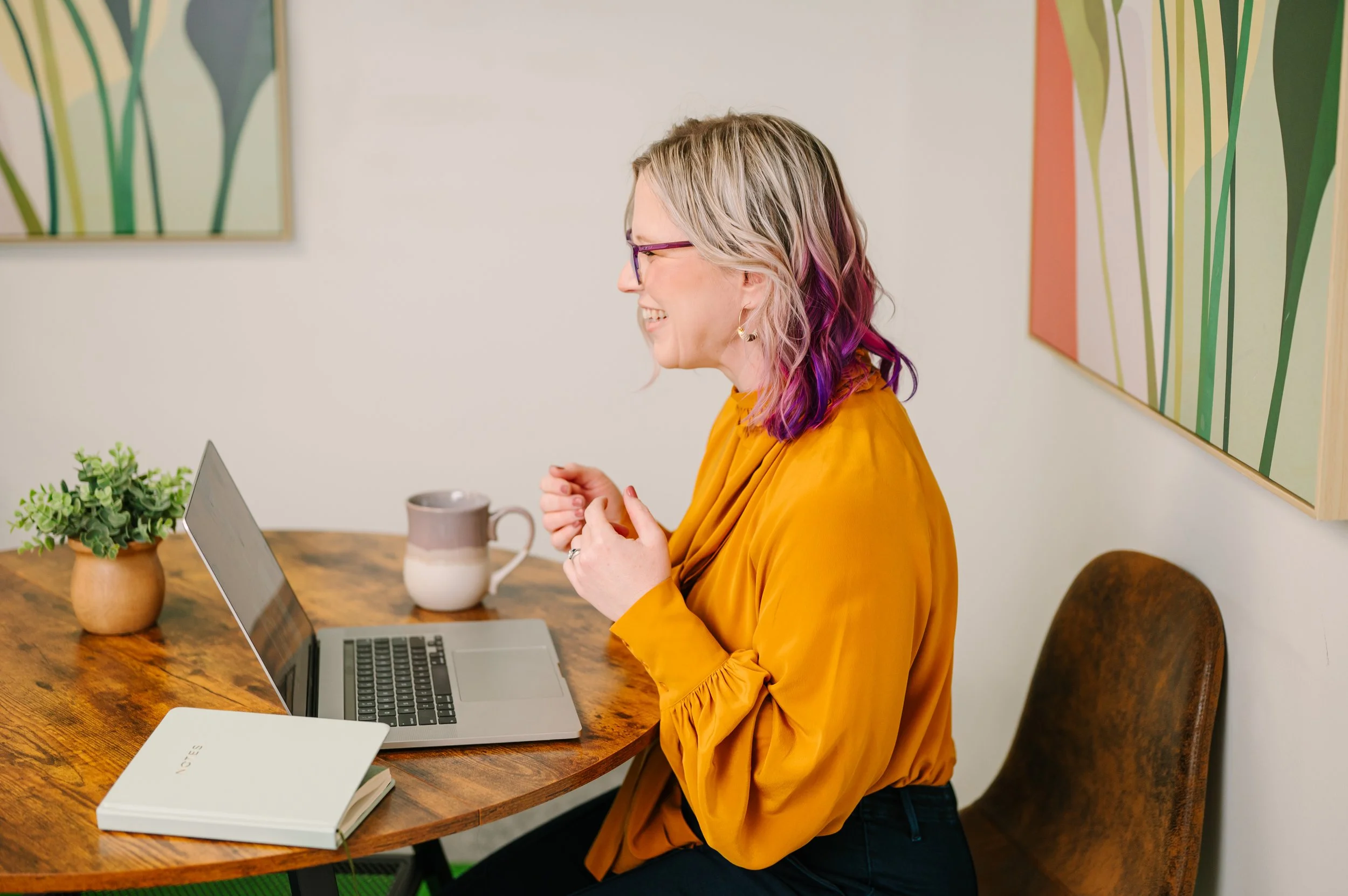 Dr. Emmie Strassberg laughing on a video call while seated at a table with a laptop and mug, wearing a mustard yellow top and purple glasses.