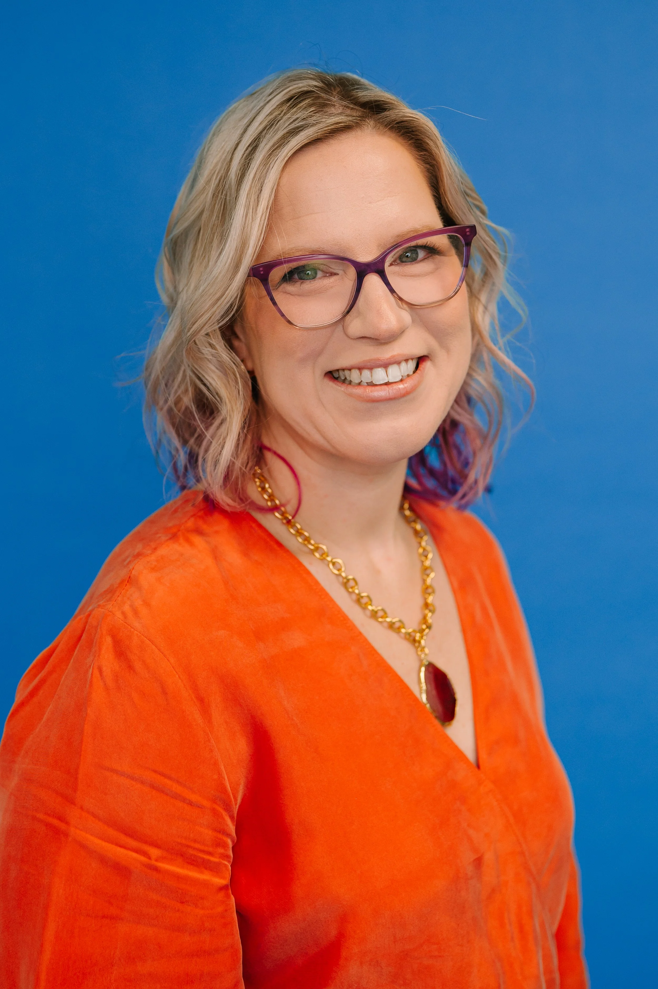 Dr. Emmie Strassberg smiling in a professional headshot, wearing an orange top and purple glasses, against a blue background.