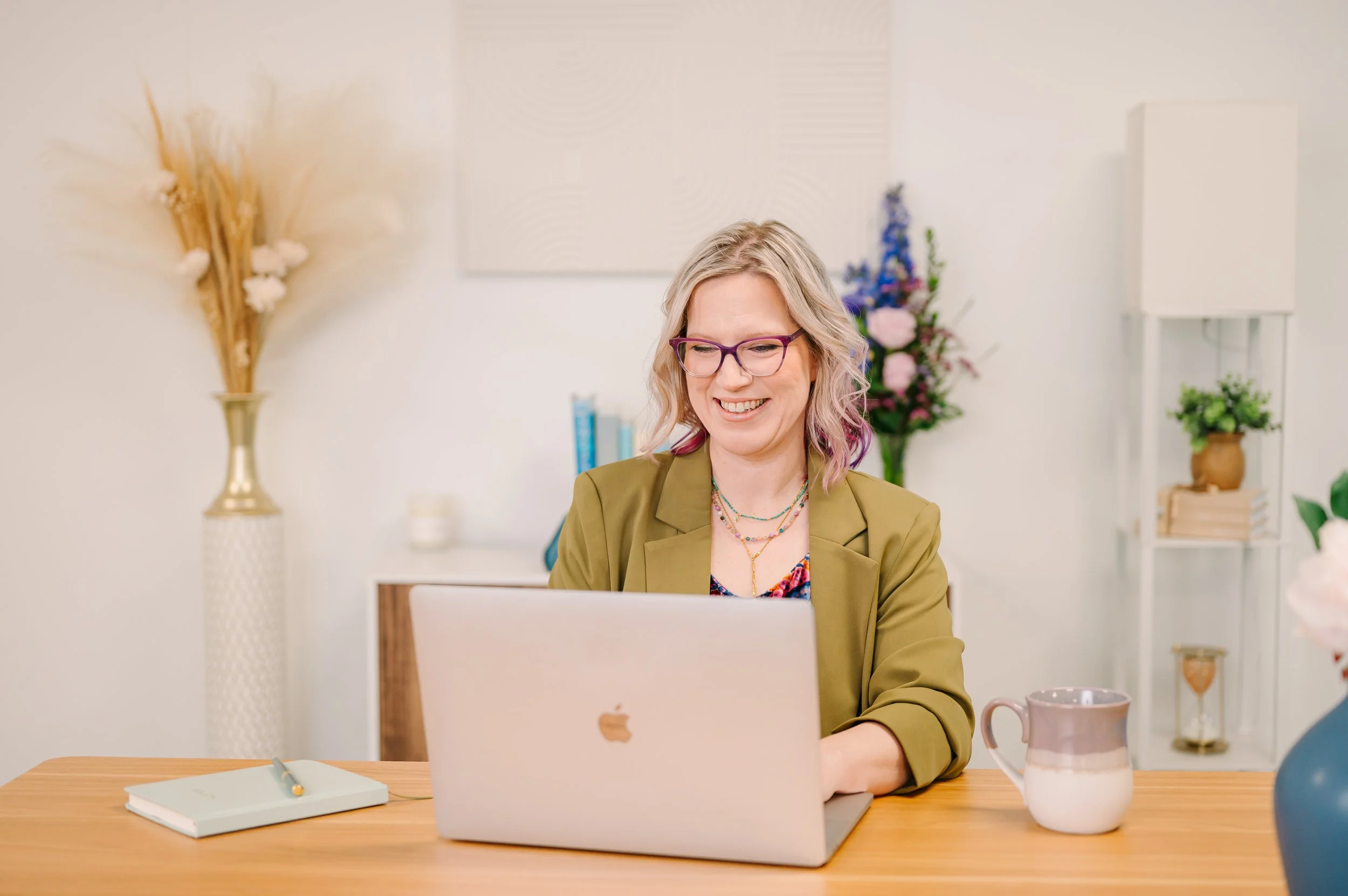 Dr. Emmie Strassberg smiling while working on a laptop, wearing an olive green blazer and purple glasses in a bright office.