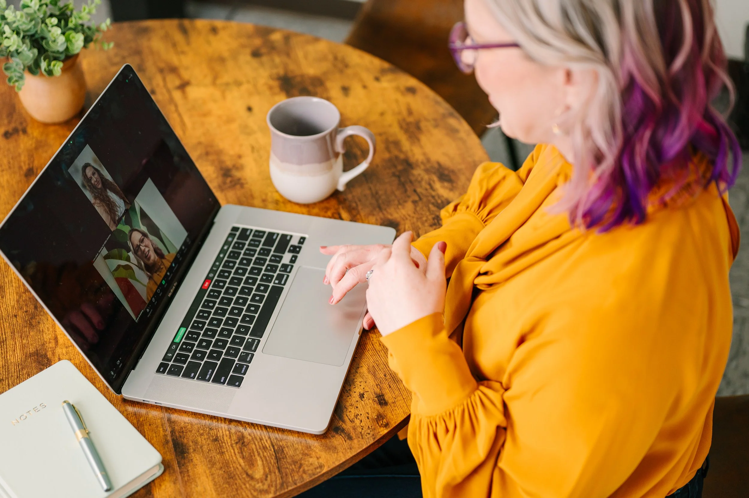 Dr. Emmie Strassberg conducting a video therapy session on a laptop at a wooden table, with a notebook and mug nearby.