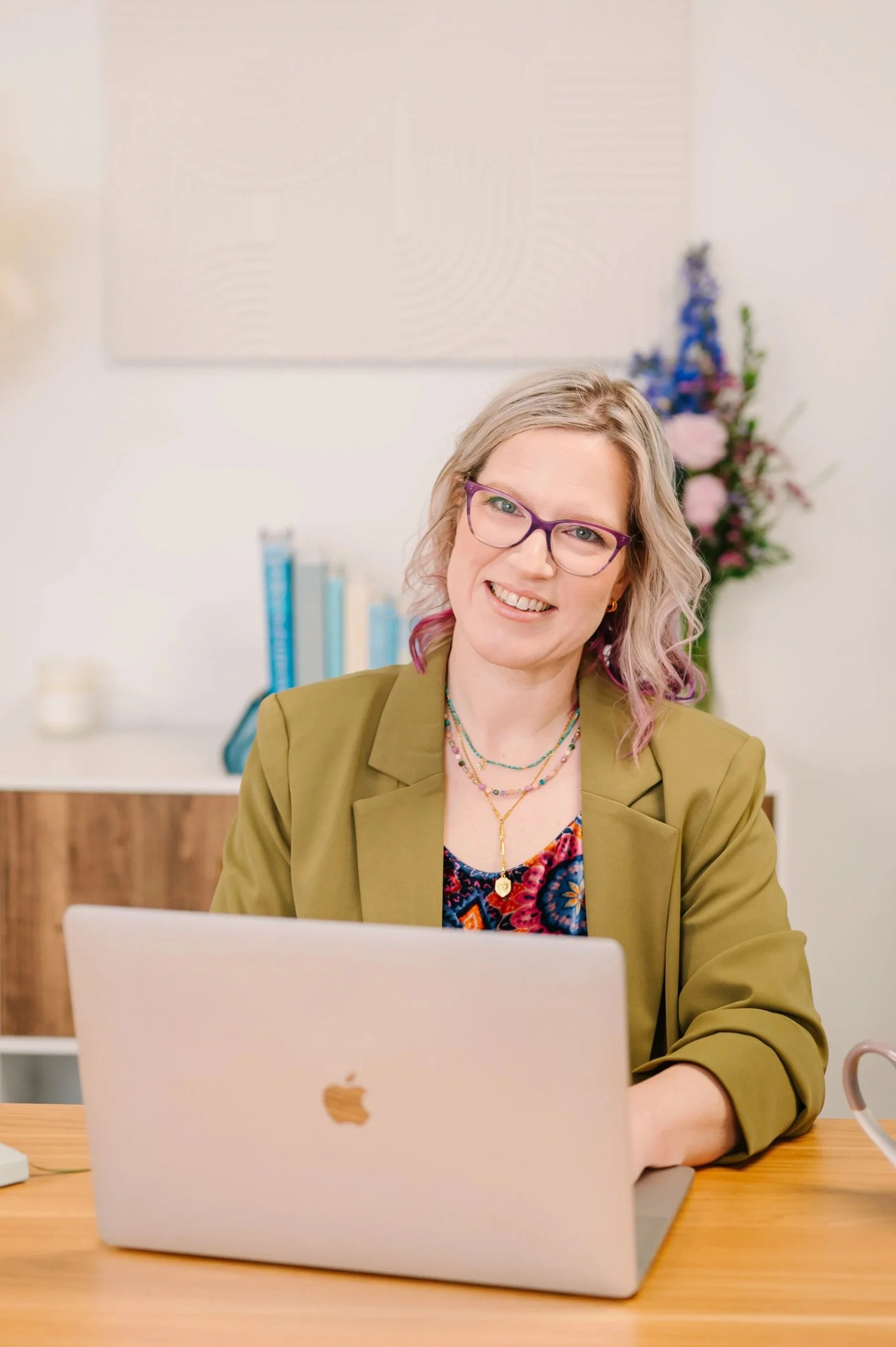 Dr. Emmie Strassberg smiling at her desk with a laptop, wearing an olive green blazer and purple glasses in a bright, welcoming office.