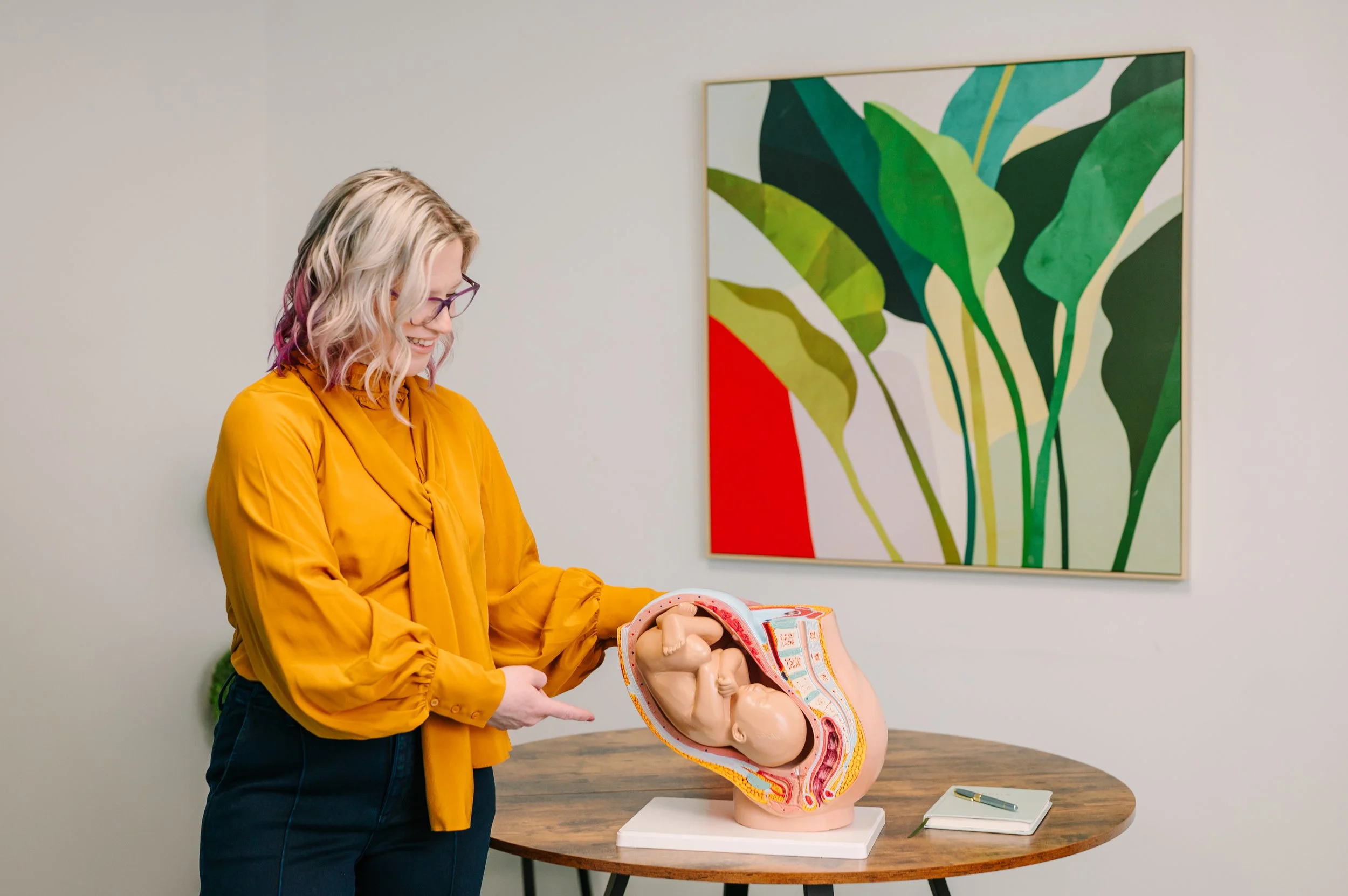 Dr. Emmie Strassberg presenting an anatomical pregnancy model, wearing a mustard yellow top, with a colorful botanical painting in the background.