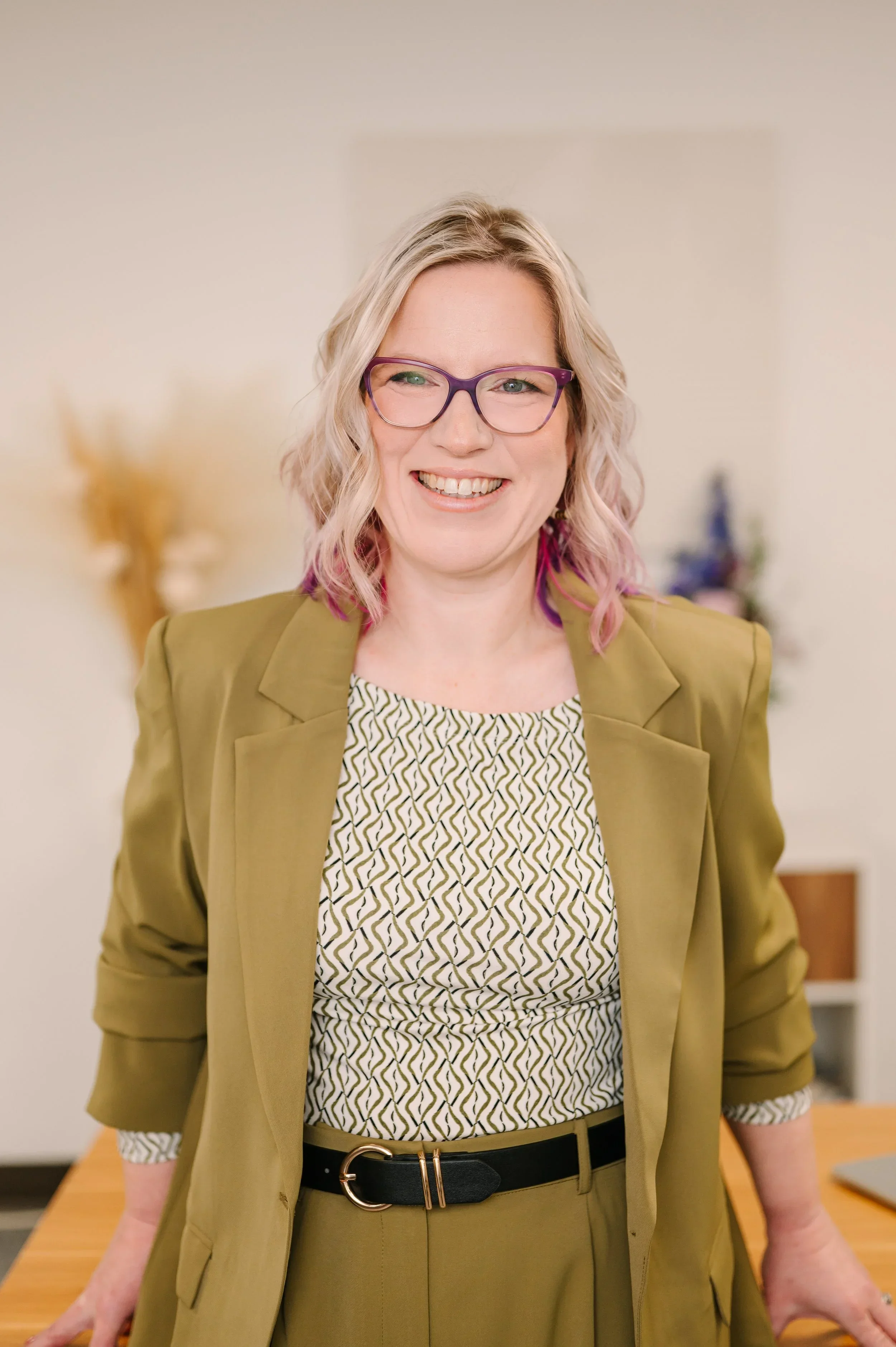 Dr. Emmie Strassberg smiling in a professional headshot, wearing an olive green blazer and purple glasses.