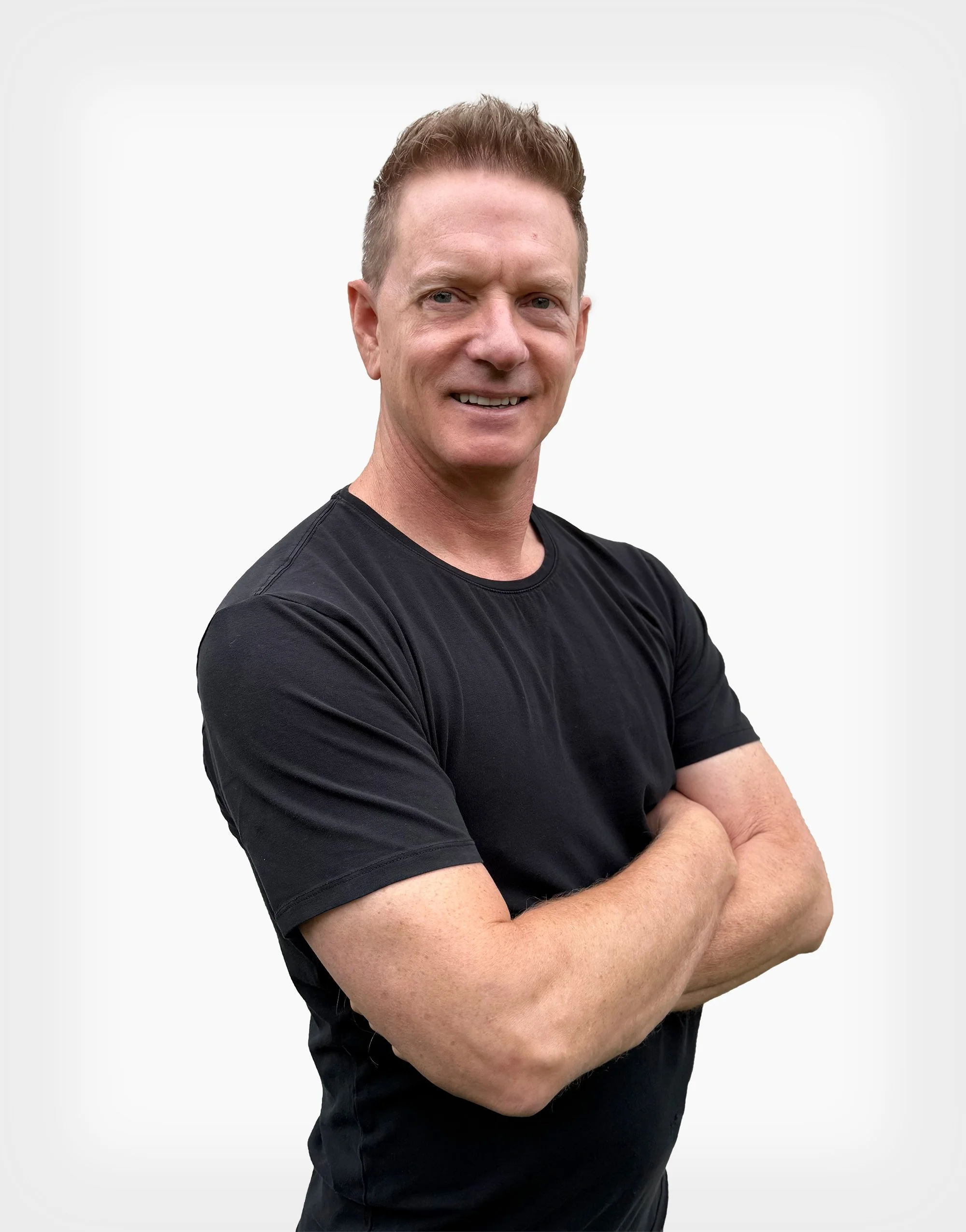 A man with short, brown hair, wearing a black t-shirt, standing with arms crossed, smiling at the camera against a plain white background.