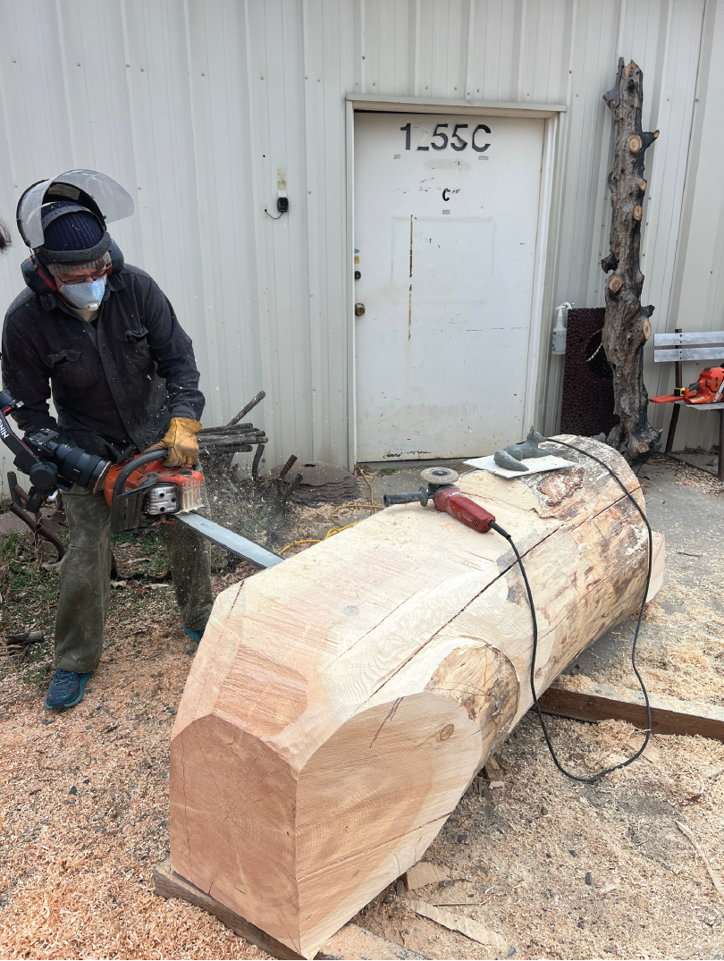 A man wearing a face mask, ear protection, and gloves is cutting a large log with a chainsaw outdoors near a white building with a gray door. Sawdust is flying as he cuts through the wood, which is lying on the ground.