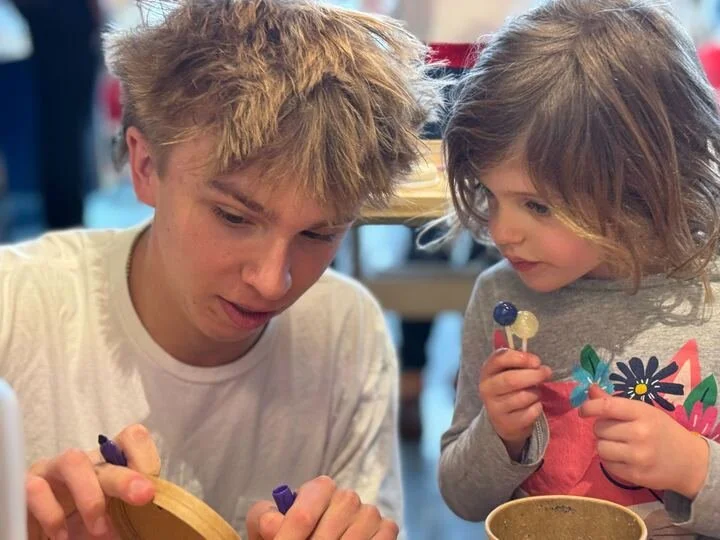 A young man with messy blonde hair and a little girl with shoulder-length brown hair are looking at a craft project together. They are in an indoor setting, possibly a cafe or craft studio, with colorful background elements.