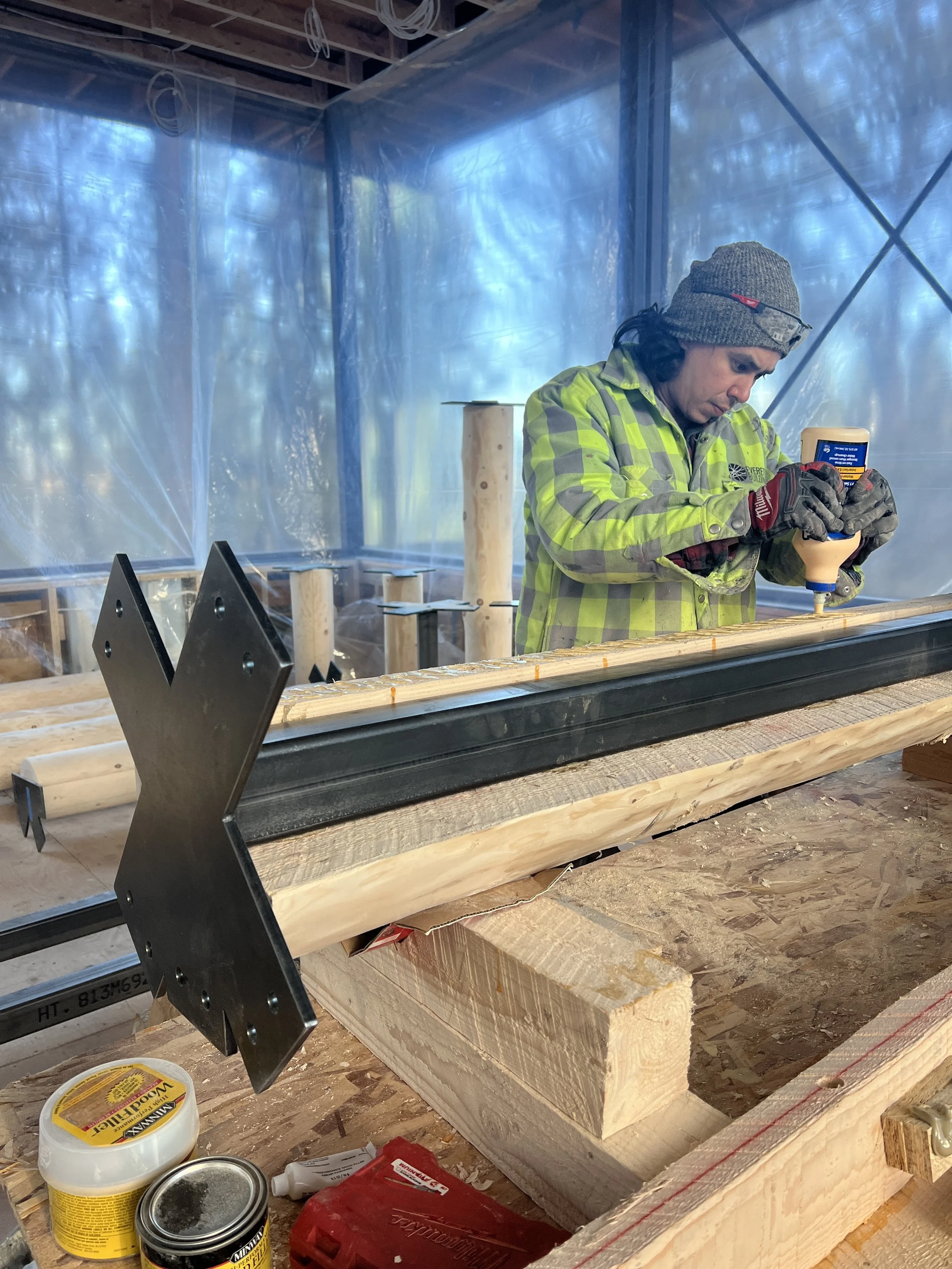 A man wearing a gray beanie, yellow and gray checkered jacket, and gloves is working with construction materials inside a partially built structure, applying adhesive to a wooden beam.