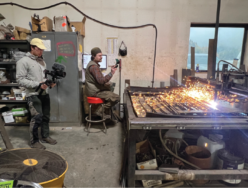 Two men are working in a welding shop, one operating a welding machine with sparks flying, and the other filming with a camera.