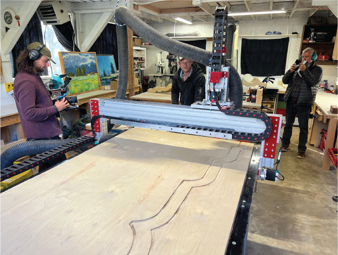 Two people using a CNC machine to carve patterns into a large wooden board, with one person recording the process and another taking photos, in a woodworking workshop.