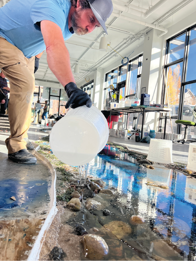 A man with a gray hat, blue shirt, and tan pants is pouring water from a white container into a small indoor stream with rocks, reflecting sunlight through large windows.