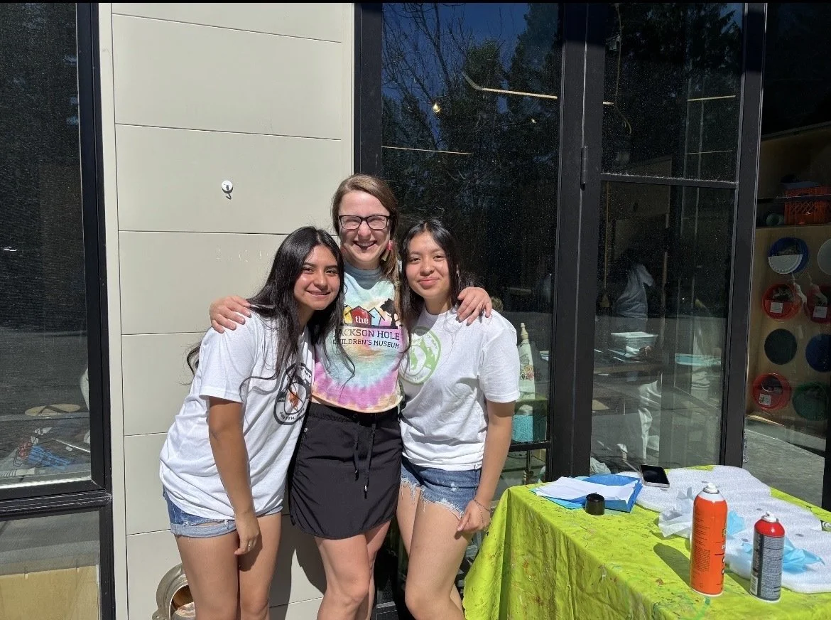 Three young women standing together outside a building, smiling at the camera, with a table of supplies covered by a yellow tablecloth nearby.