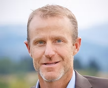 A smiling middle-aged man with short light brown hair and blue eyes, wearing a suit jacket, outdoors with a blurred background of nature and mountains. Brendan McDermott Treasurer of the board of directors