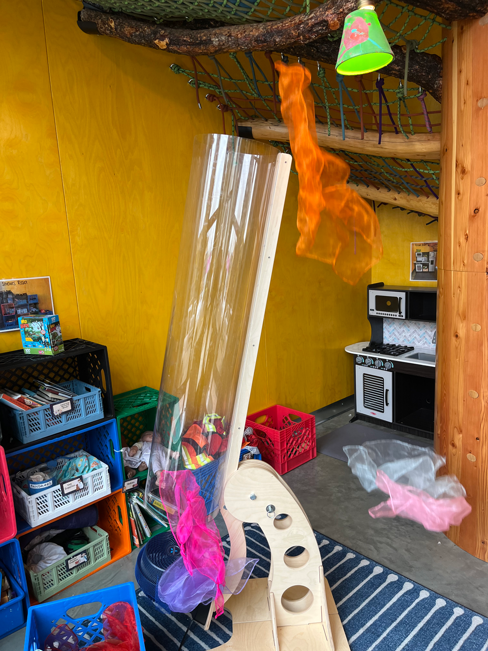 Indoor play area featuring a tree branch with a hanging paper lamp, a fire simulation on the branch, a wooden structure with holes, colorful tissue paper decorations, and a play kitchen in the background.