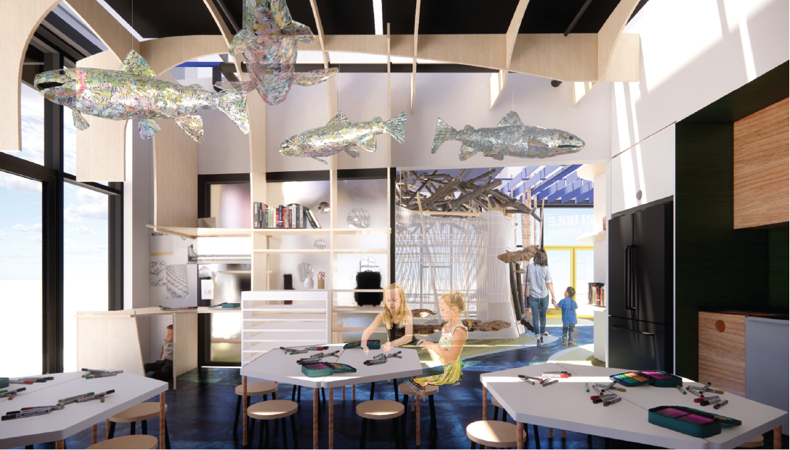 Children's play area inside a modern building with fish-shaped sculptures hanging from the ceiling, children playing and adults walking in the background, with large windows and natural light.